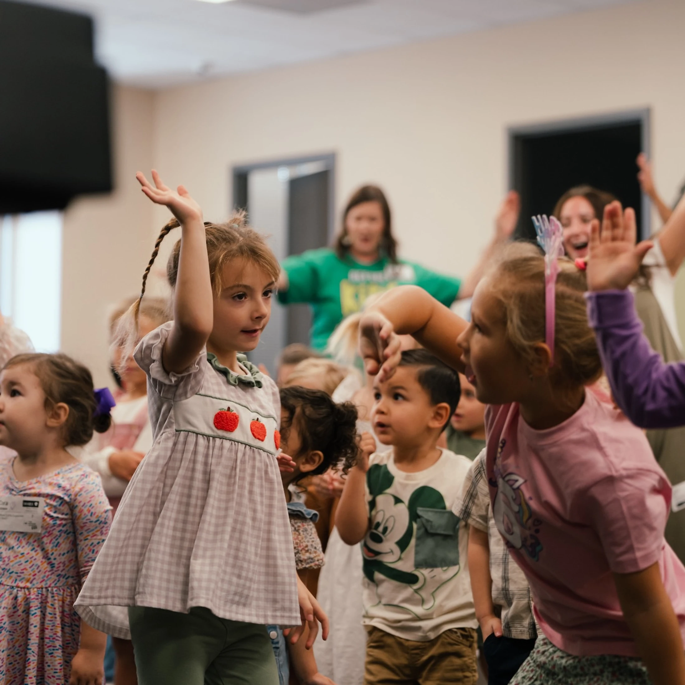 A group of young children eagerly raises their hands to participate in a fun activity in a classroom, with adults leading in the background.