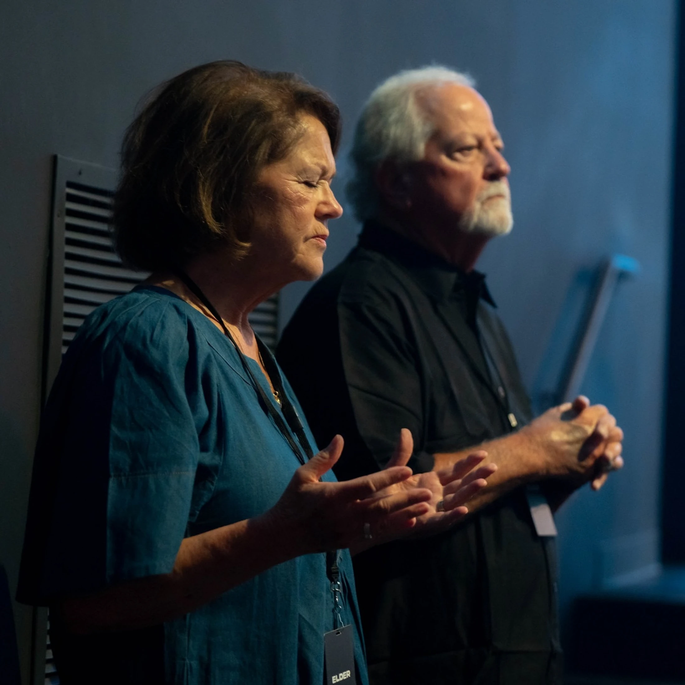 An older woman with eyes closed and an older man with a white goatee stand side-by-side with hands clasped, appearing to be in prayer or worship.