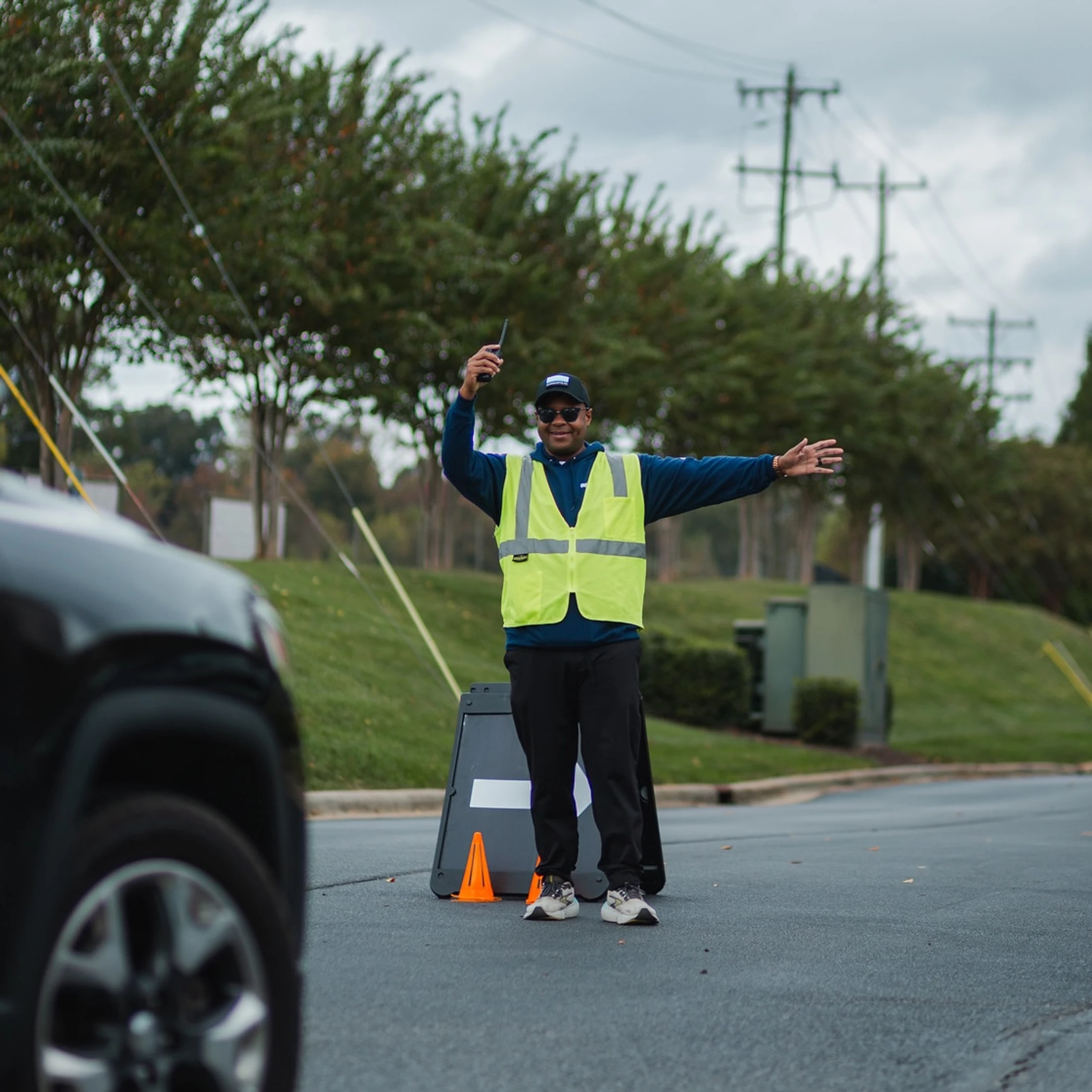 A smiling man in a yellow safety vest directs a black car in a parking lot. He holds a walkie-talkie in one hand and has his other arm outstretched.