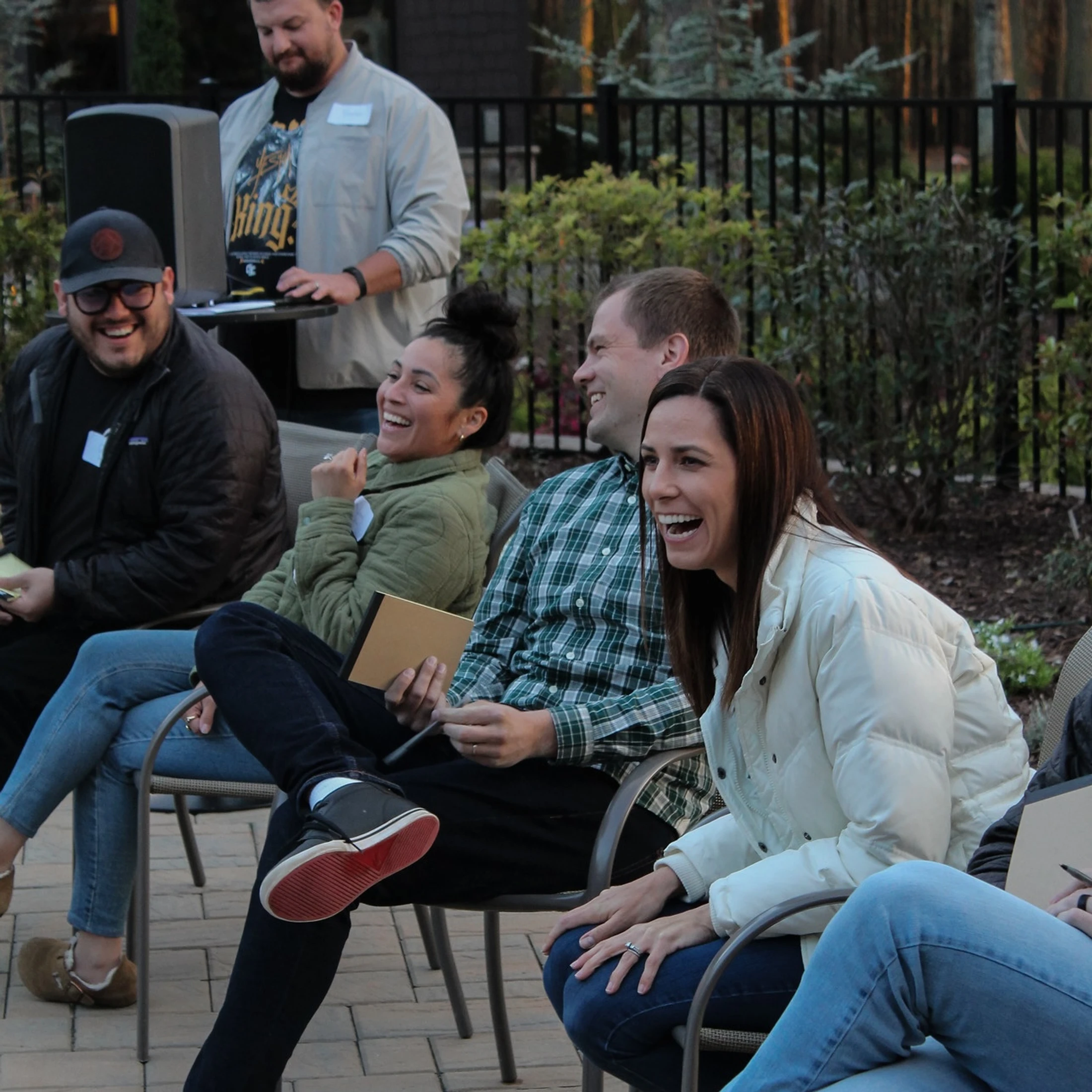 A group of four adults sitting in chairs outdoors and laughing together. In the background, a man stands behind a speaker at a small table.