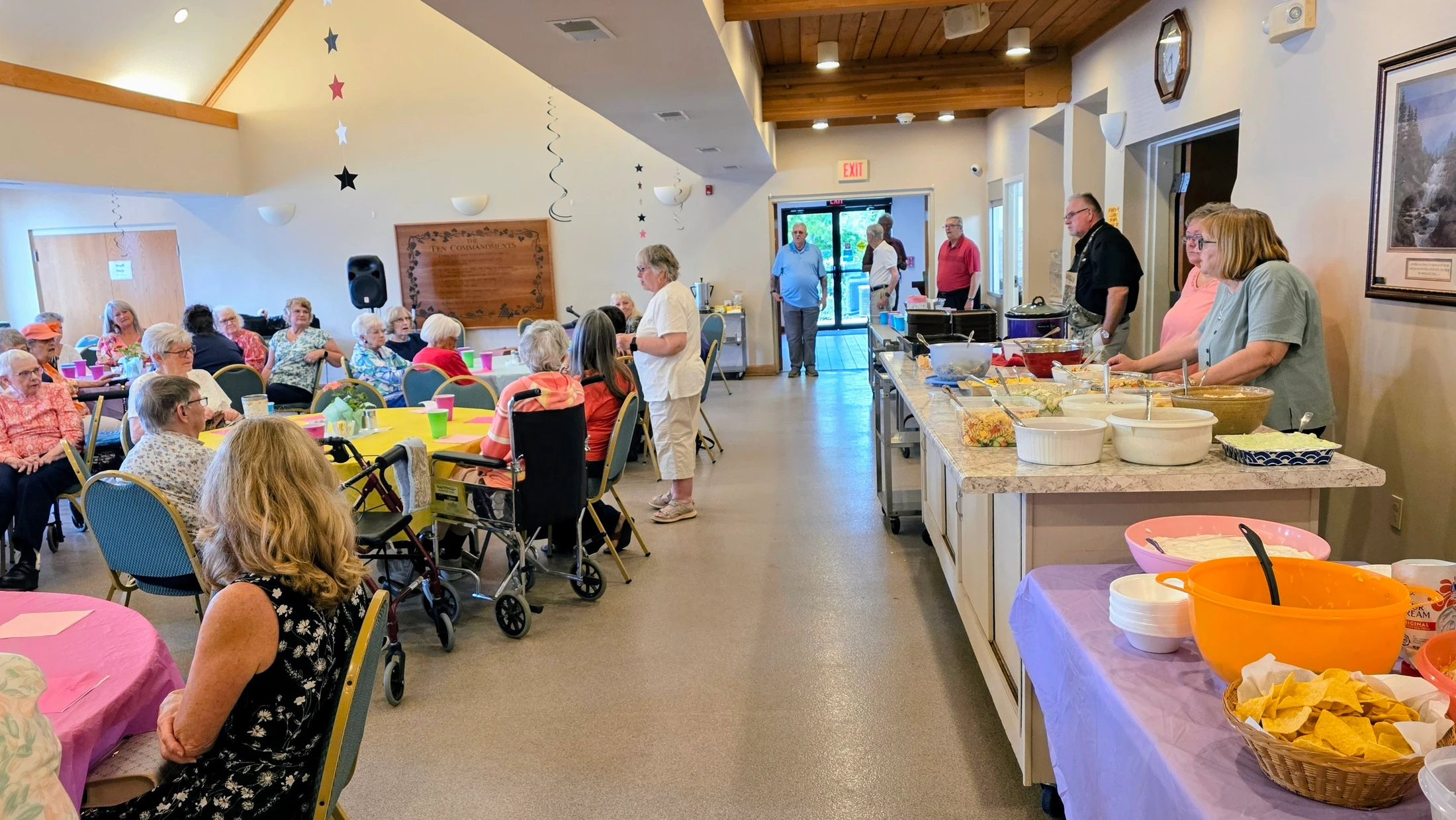 A group of seniors gather for a meal in a large room. People are seated at tables, while others line up at a buffet counter laden with various dishes.