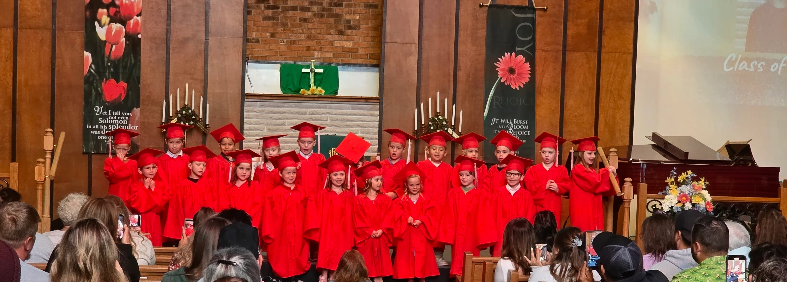 A group of young children in red graduation caps and gowns stand on a stage in a church, singing for an audience during their graduation ceremony.