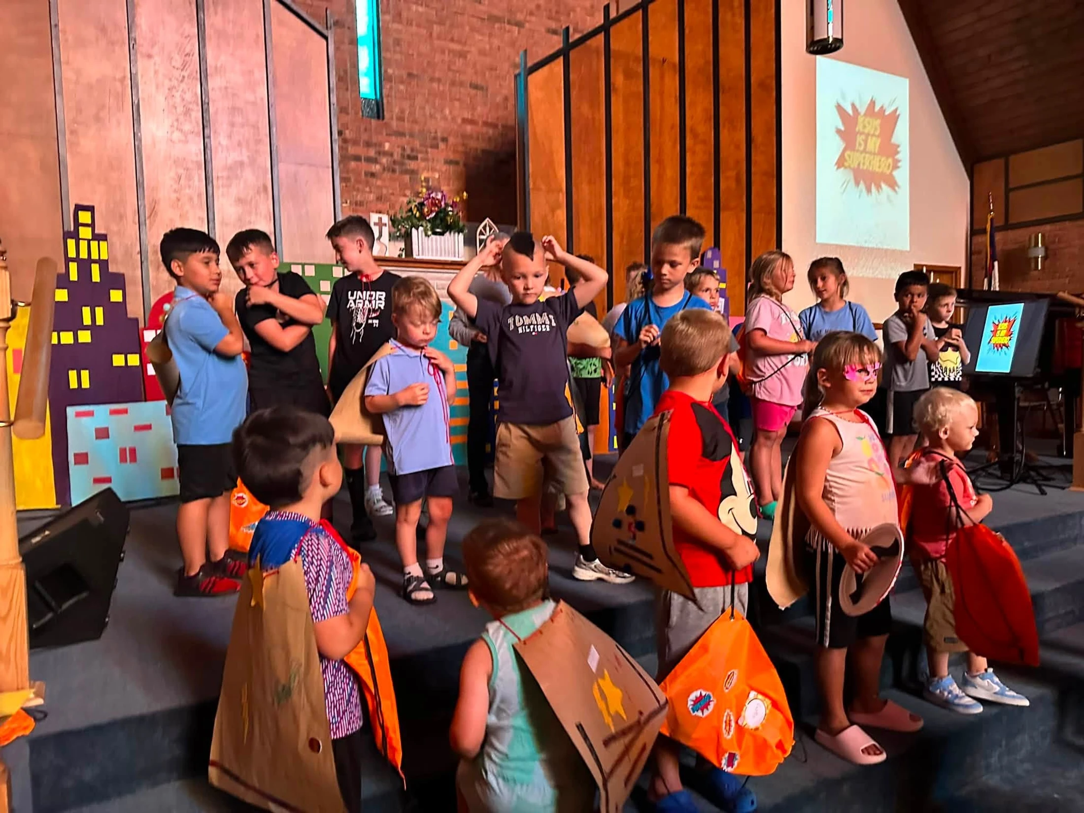 A group of children stand on a stage in a church wearing homemade superhero capes. A screen in the background reads, 'Jesus is My Superhero'.