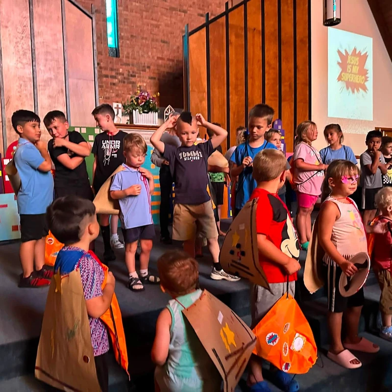 A group of children stand on a stage in a church wearing homemade superhero capes. A screen in the background reads, 'Jesus is My Superhero'.