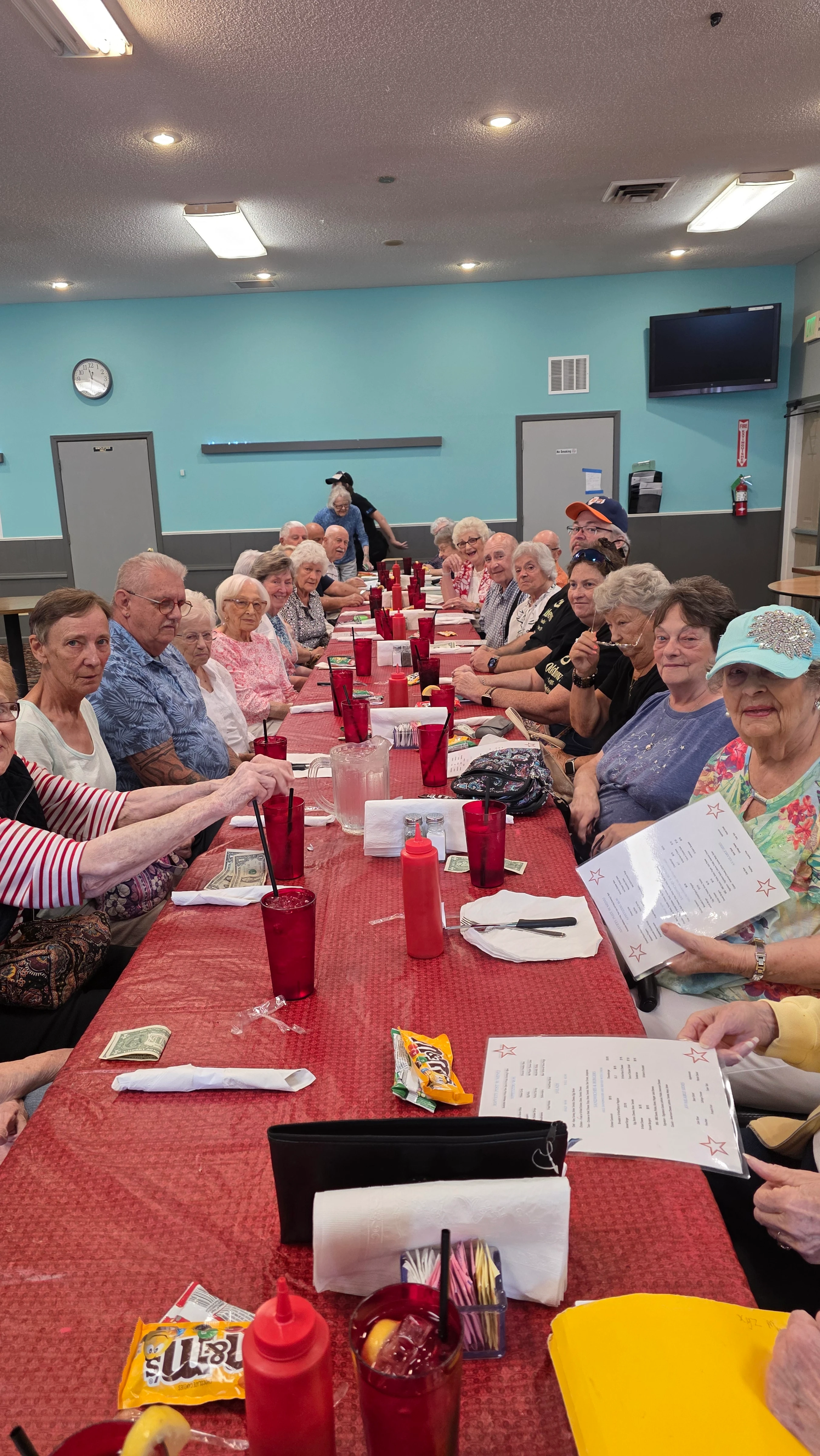 A large group of seniors sits at a long dining table with a red tablecloth in a community hall, smiling and looking at menus before their meal.