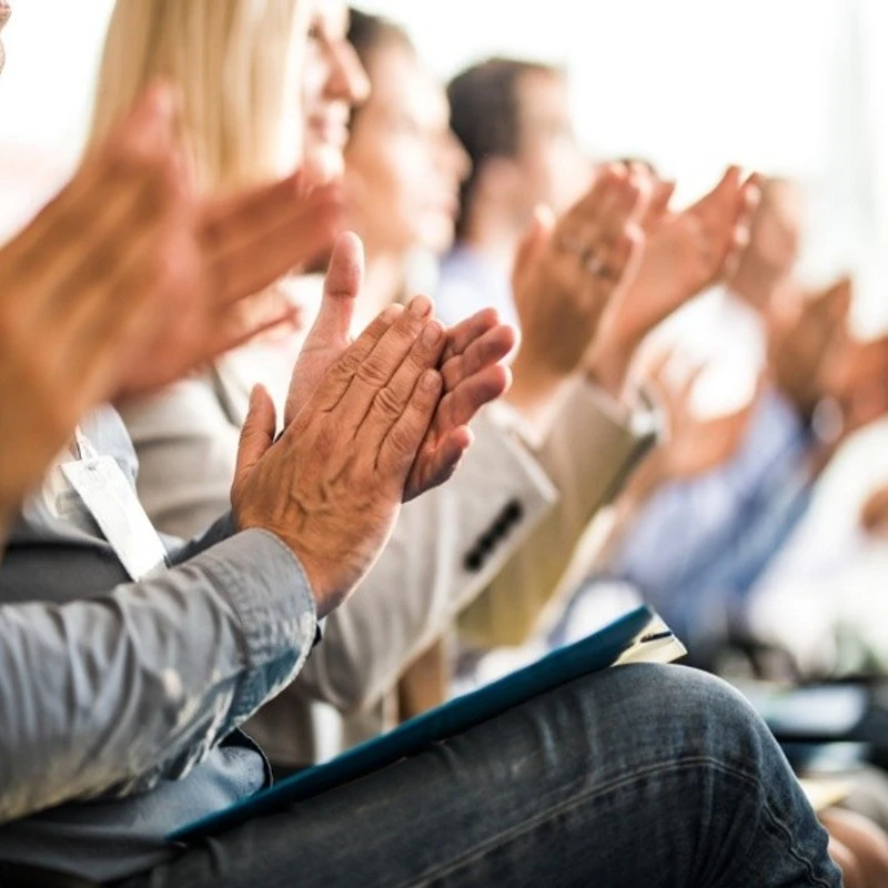 A row of people in a business setting clapping, with a close-up focus on the hands of a man in the foreground. The background is softly blurred.