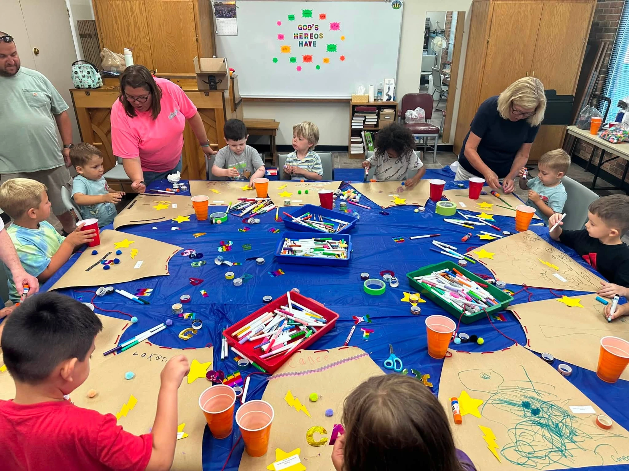 A group of young children and adults sit around a long blue table, decorating paper superhero capes with markers, stickers, and other craft supplies.