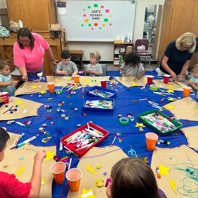 A group of young children and adults sit around a long blue table, decorating paper superhero capes with markers, stickers, and other craft supplies.