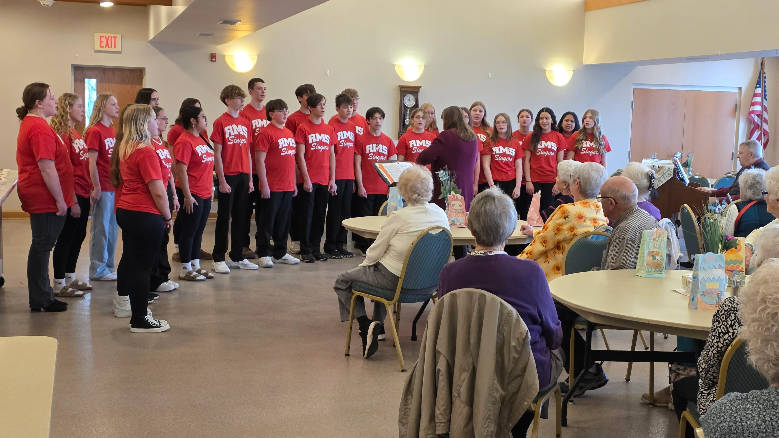 A youth choir wearing matching red t-shirts performs for an audience of senior citizens seated at round tables in a brightly lit community hall.