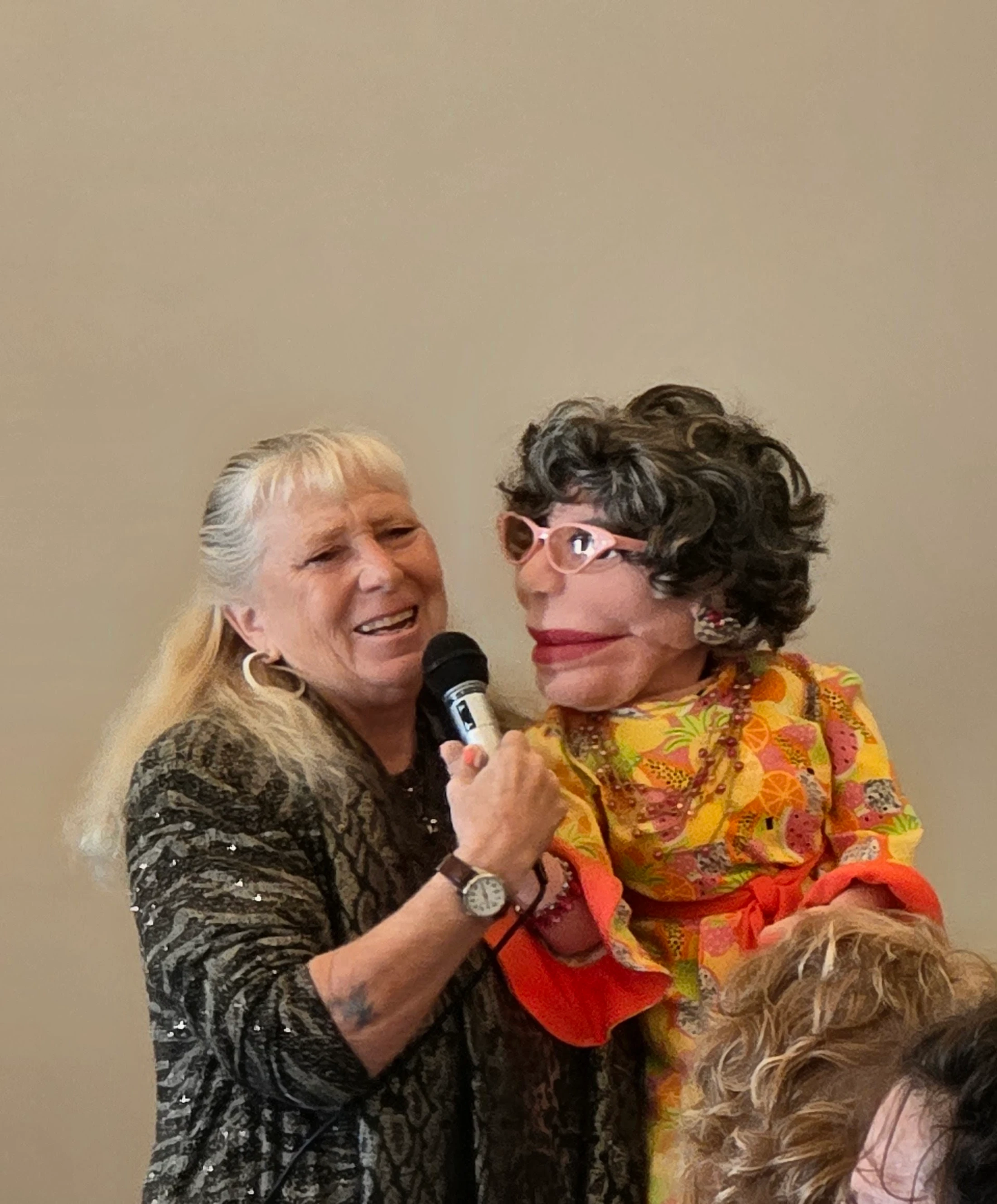 A smiling woman with long graying-blonde hair holds a microphone to a puppet of an older woman with curly gray hair and pink cat-eye glasses.