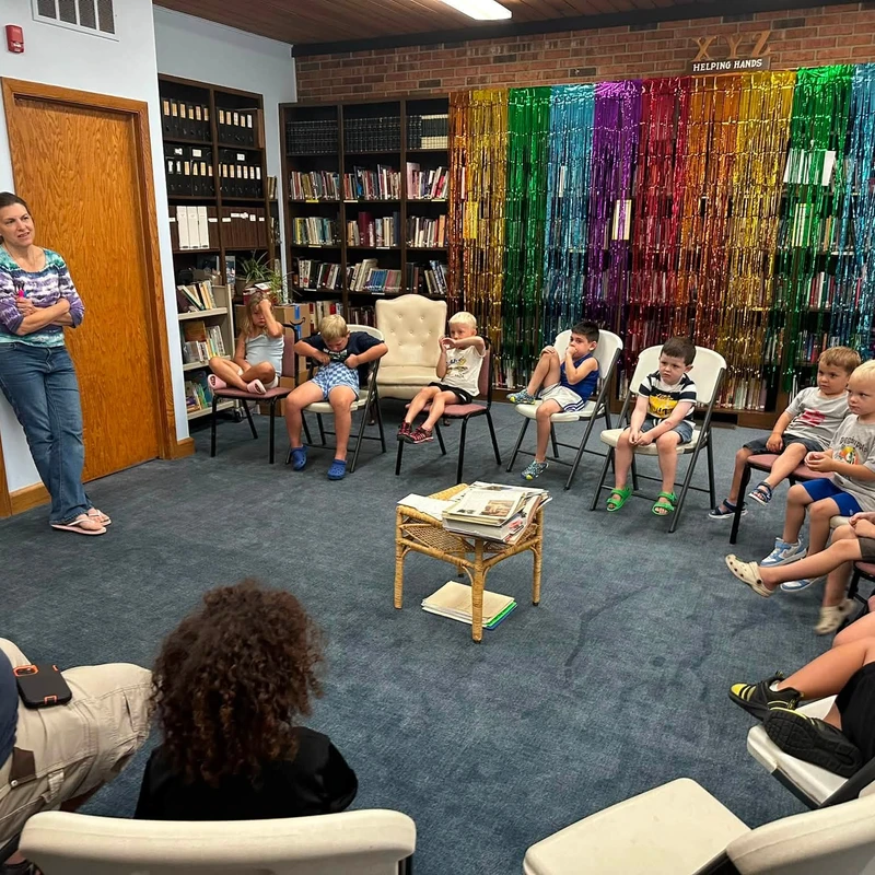A group of young children sit in chairs in a semi-circle, listening to a woman standing in a library room with a colorful rainbow backdrop.
