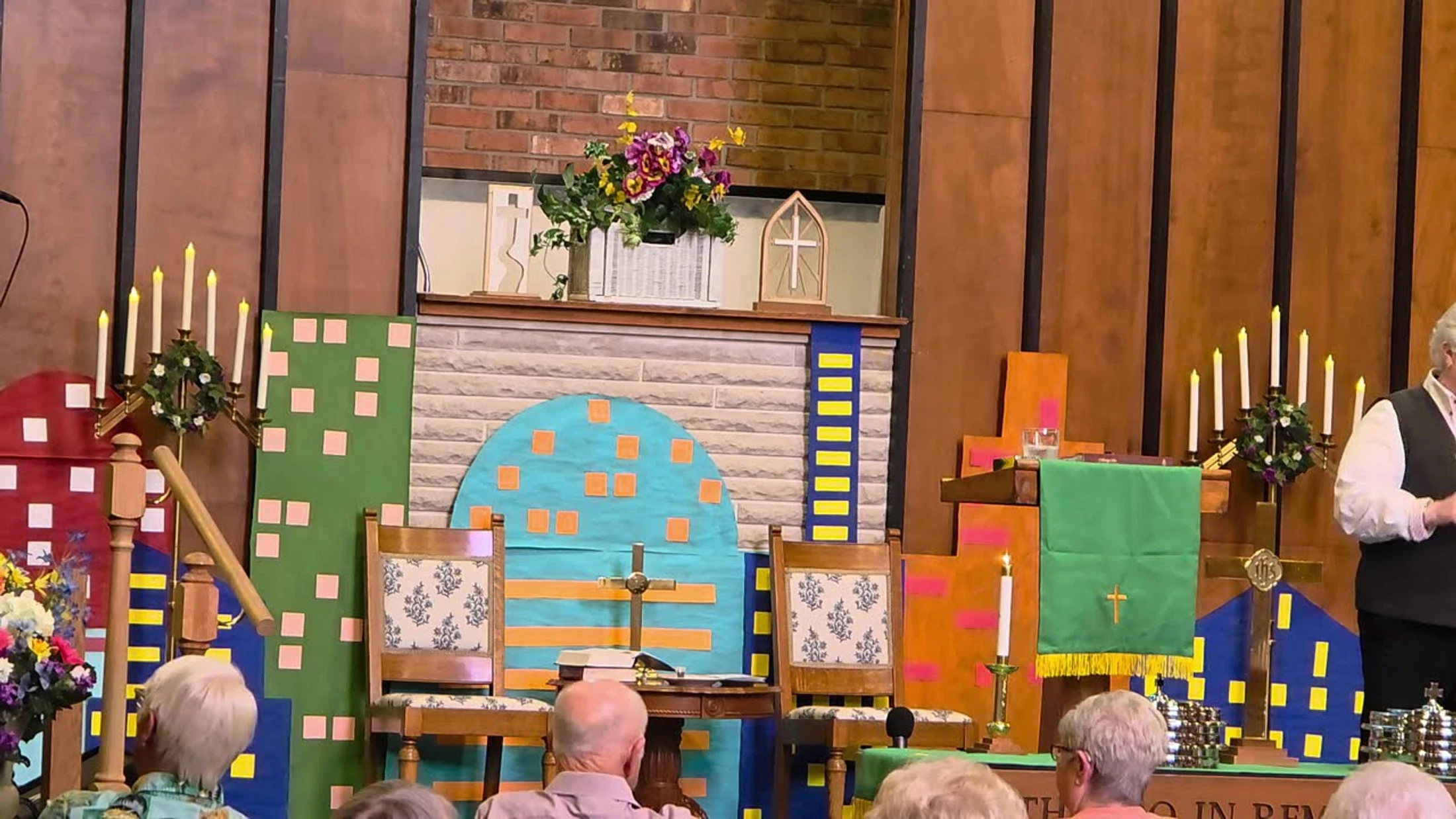 A church altar decorated with colorful cutouts of city buildings for a service. A congregation sits in the foreground, facing the decorated chancel.
