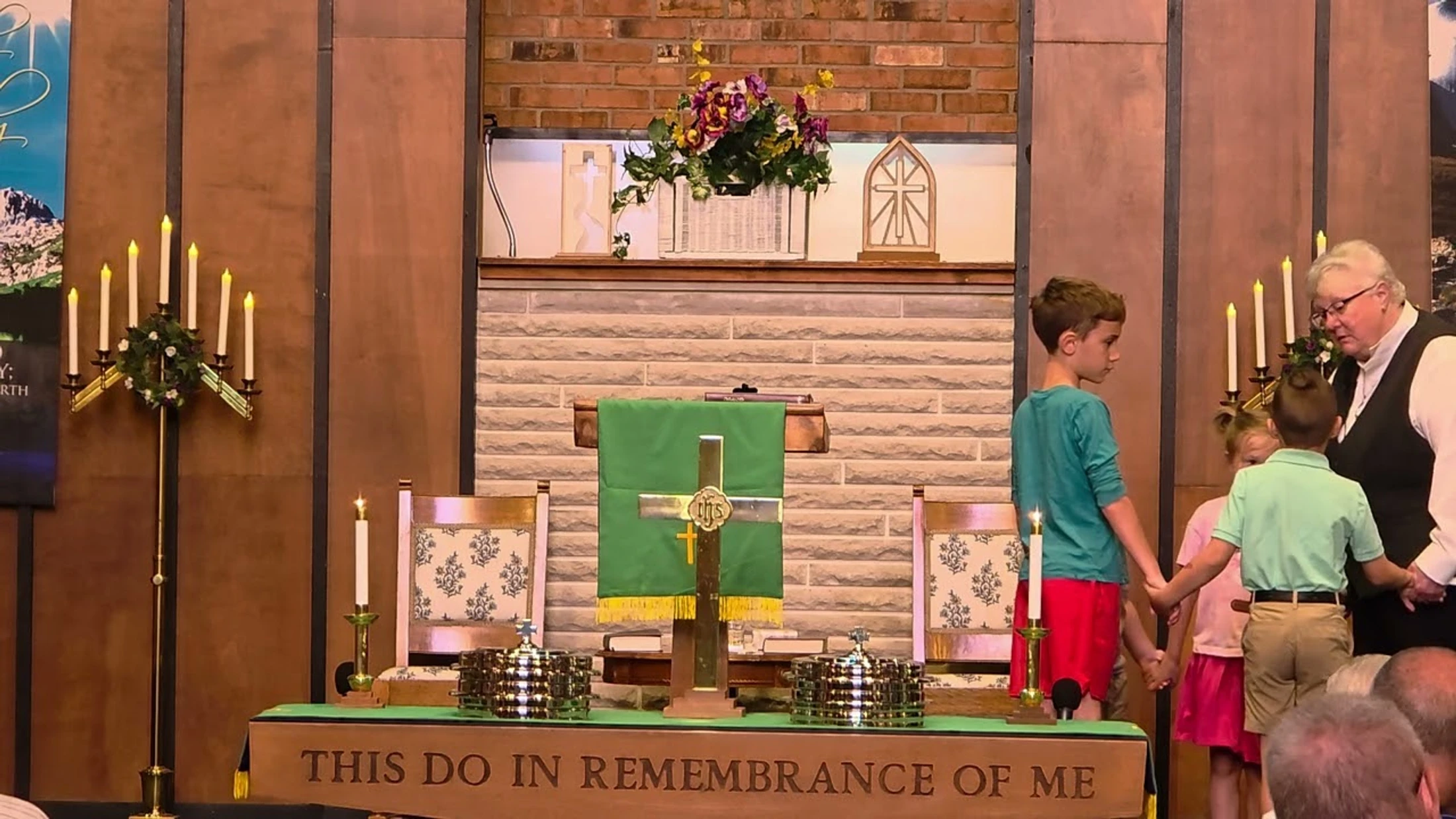 A female pastor in a vestment holds hands with three young children in a line at the front of a church, standing near the altar during a service.