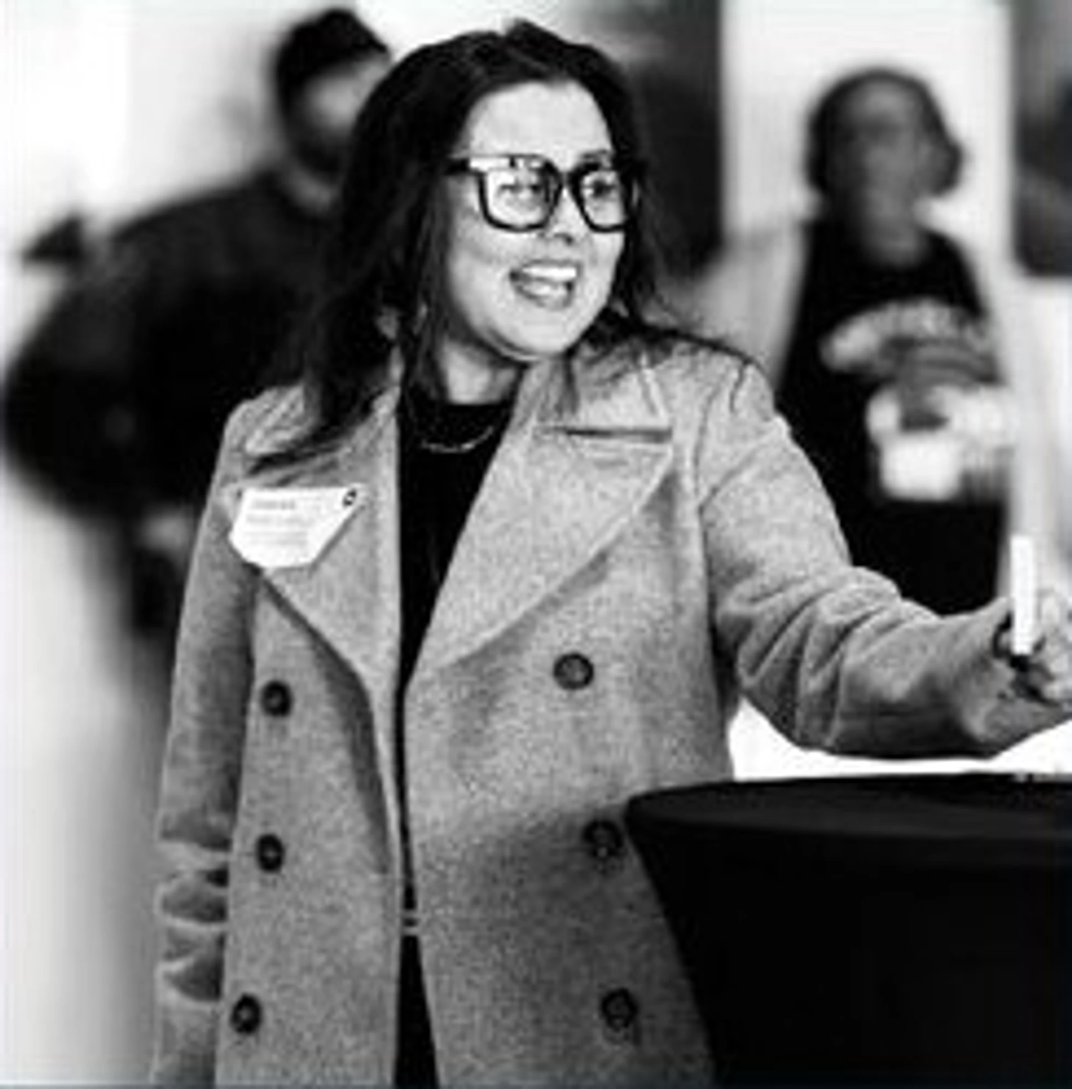 A black and white photo of a smiling woman with dark hair and large glasses. She wears a pea coat with a name tag and stands at a table during an event.