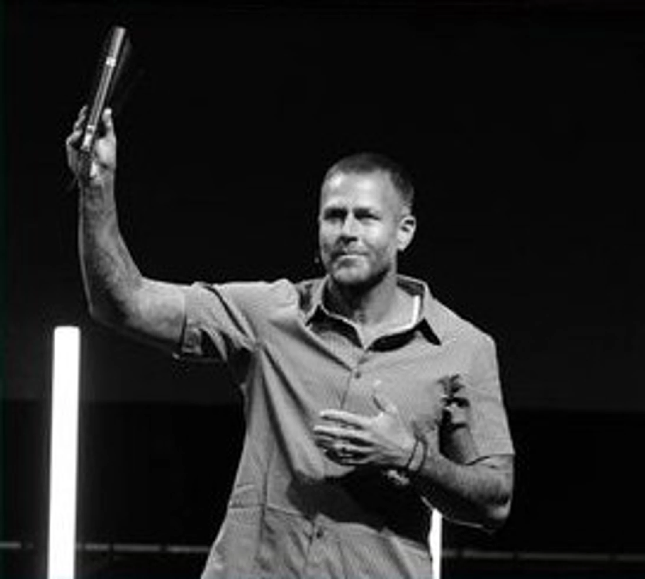 A black and white photo of a man in a short-sleeved shirt on stage, holding a microphone up in one hand and placing the other over his heart.