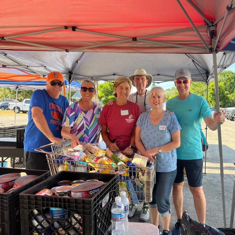 A group of six smiling volunteers stands under a red pop-up tent, posing behind a shopping cart and crates filled with donated food items.