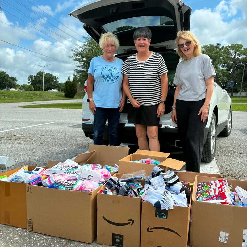 Three smiling women stand in a parking lot behind an open SUV, with several cardboard boxes filled with new children's socks and underwear in front of them.