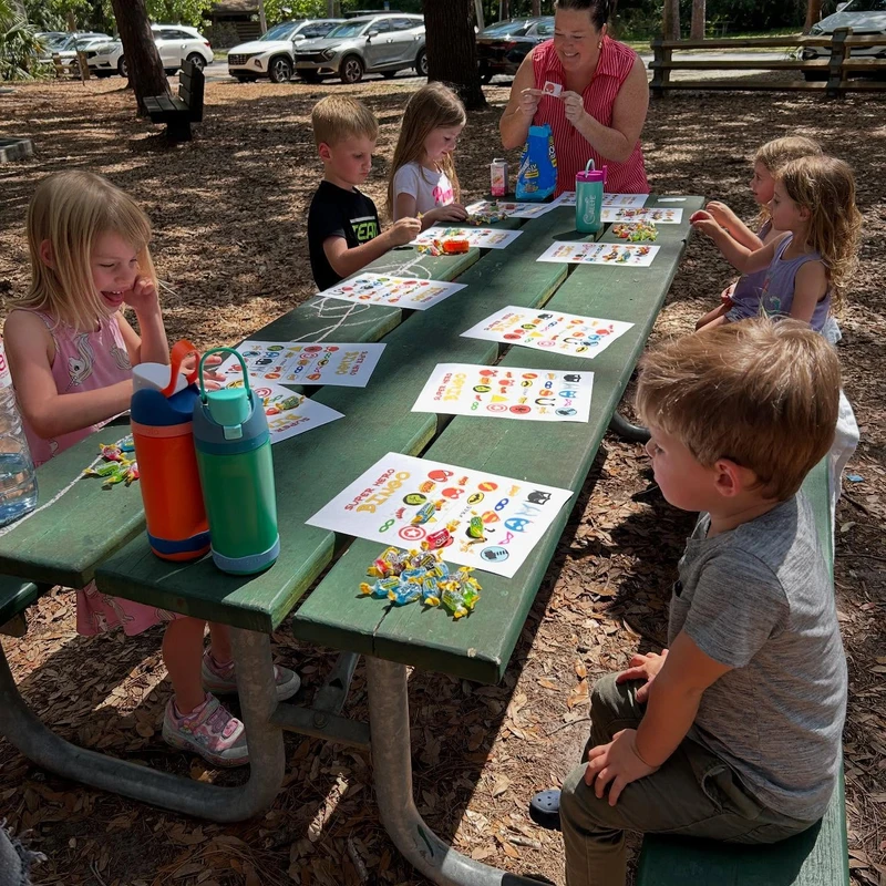 A woman and several young children sit at a long green picnic table outdoors, playing Super Hero Bingo with colorful candies as markers.