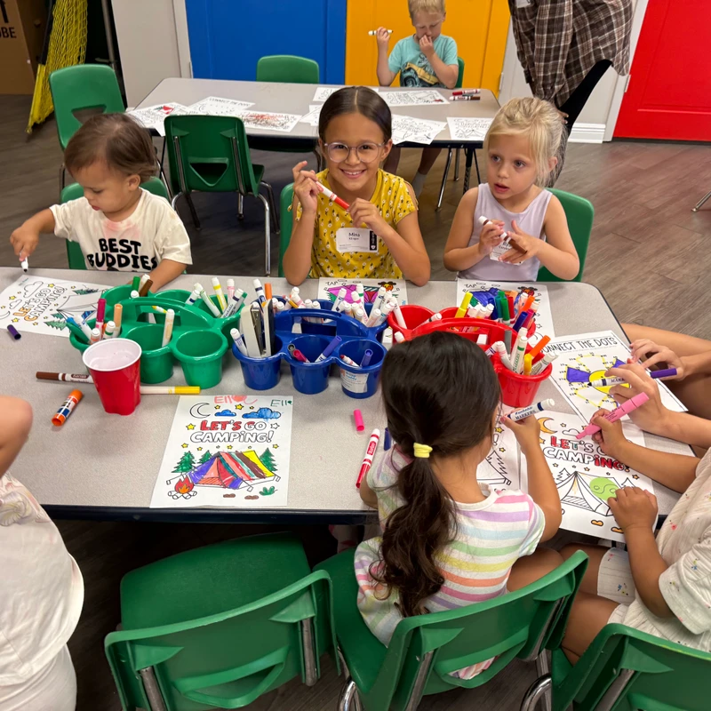 A group of young children sit around a table in a colorful classroom, using markers to color in camping-themed worksheets. One girl in a yellow shirt smiles.