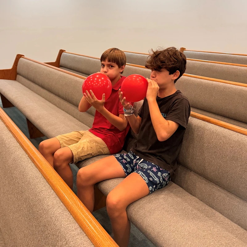 Two young boys sit side-by-side on a gray cushioned pew, each holding and blowing up a bright red balloon.