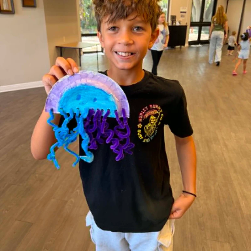 A smiling boy with brown hair holds up a colorful jellyfish craft made from a paper plate and blue and purple pipe cleaners in a large room.