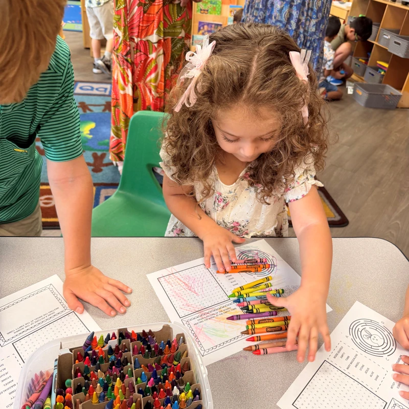 A young girl with curly hair and a pink bow sits at a table coloring on a worksheet. A large tub of colorful crayons is next to her in the classroom.