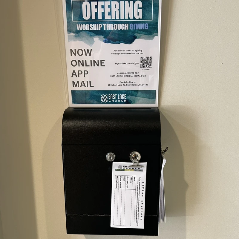 A black offering box on a wall at East Lake Church. A sign above lists giving options like online, app, or mail, and offering envelopes are attached to the box.