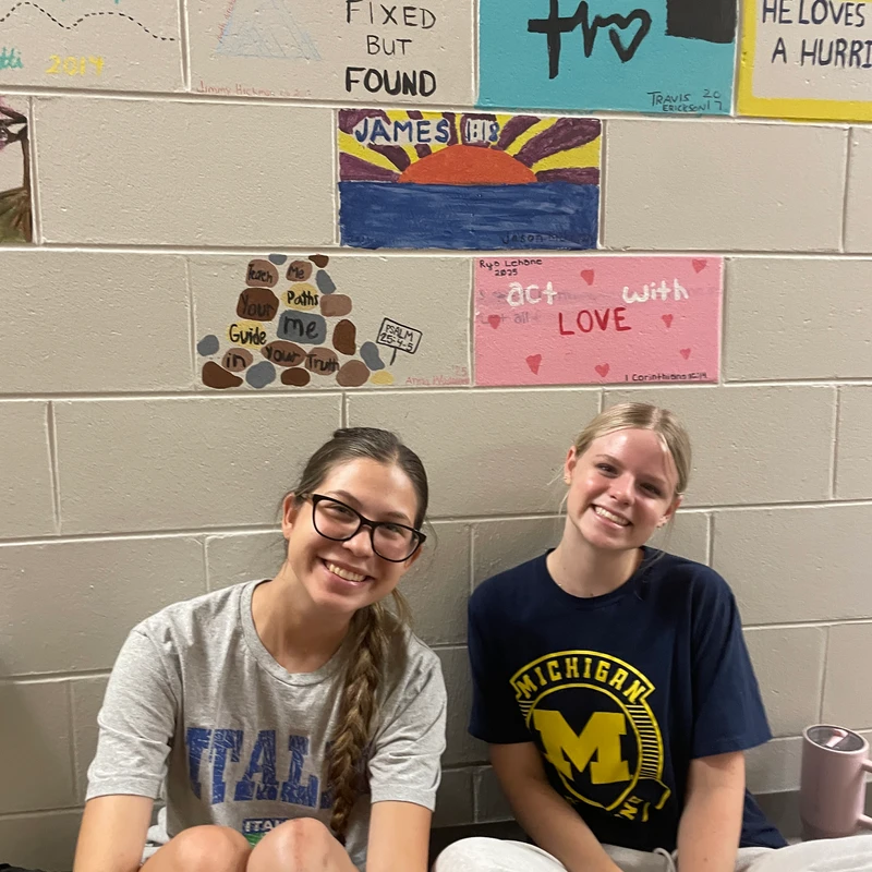 Two young women sit on the floor smiling in front of a cinder block wall decorated with small, colorful paintings containing inspirational messages.