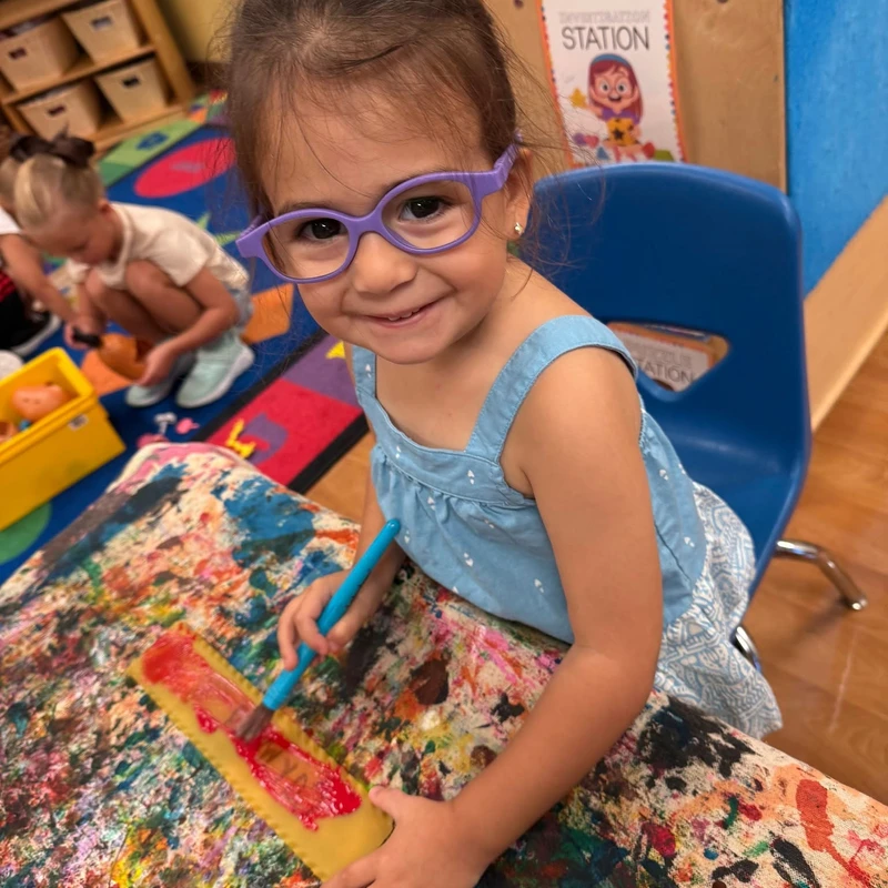 A young girl with purple glasses smiles at the camera while sitting at a messy, paint-covered table. She is painting a piece of lasagna noodle red.