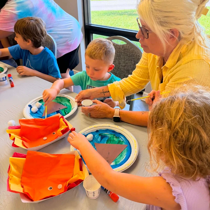 A woman with blonde hair helps three young children paint and create crafts out of paper plates and construction paper at a table.