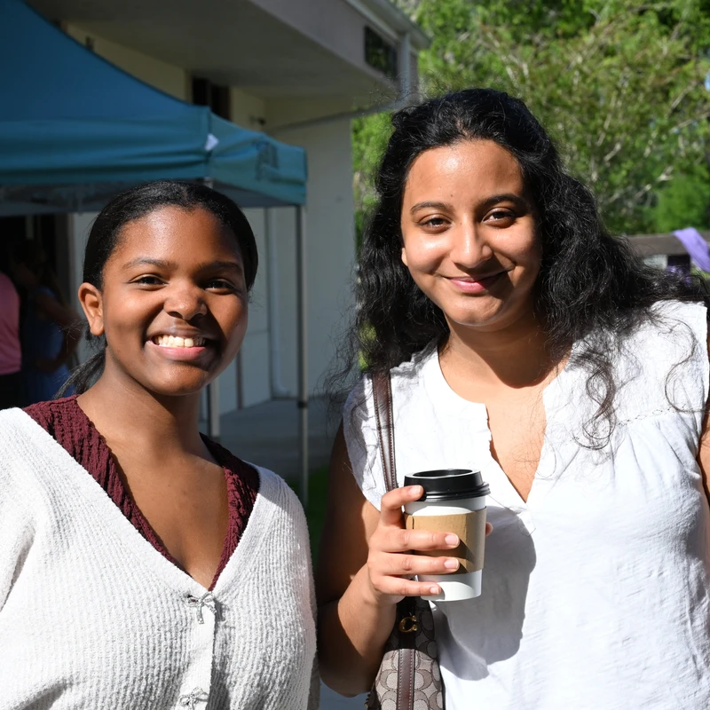 Two smiling women pose for a photo outdoors. One holds a coffee cup. In the background, a blue tent for East Lake Church and a wooden cross are visible.