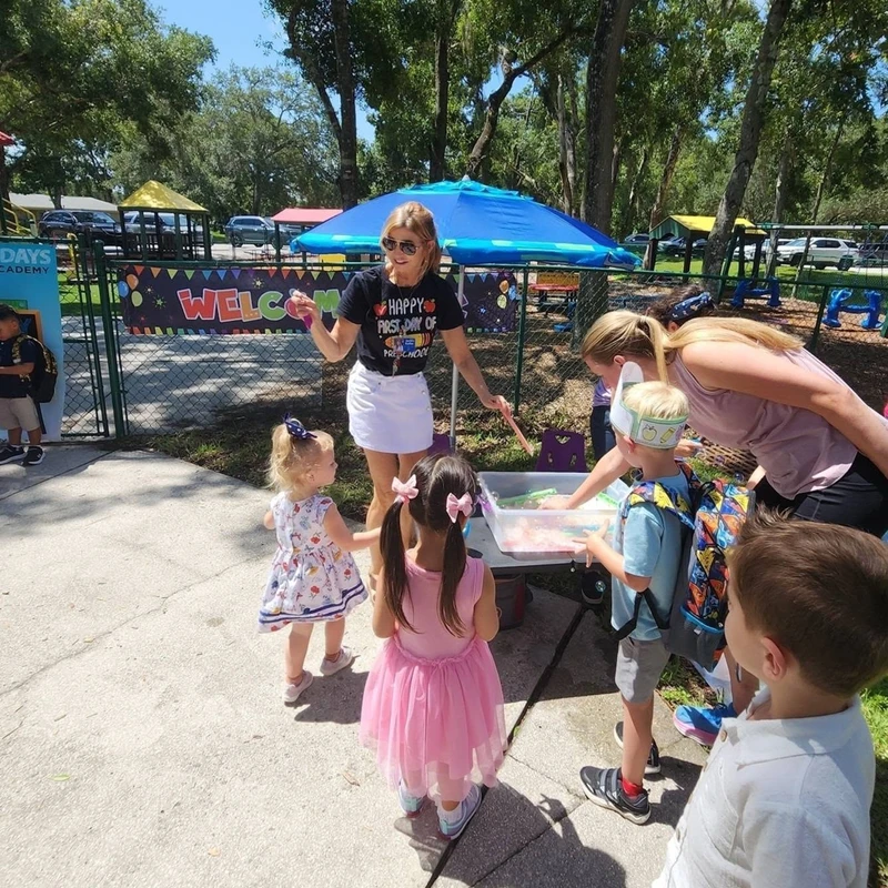 A teacher wearing a 'Happy First Day of Preschool' shirt welcomes young children outdoors as they gather around an activity table under a blue umbrella.