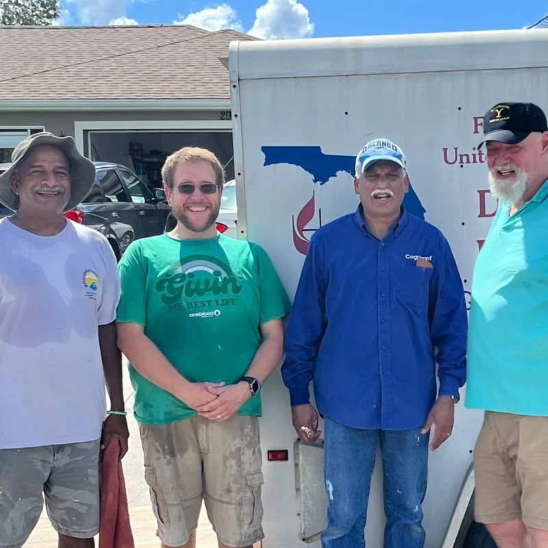 Four smiling men stand side-by-side for a group photo outdoors in front of a large white trailer and a house on a sunny day.