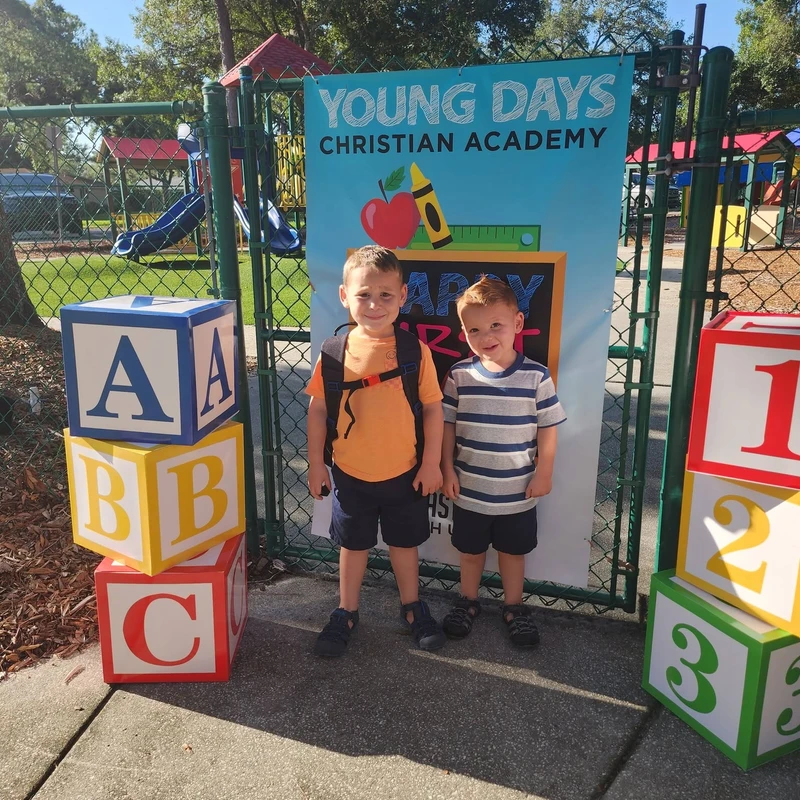 Two young boys smiling on their first day of school in front of a sign for their academy, flanked by large alphabet and number blocks.