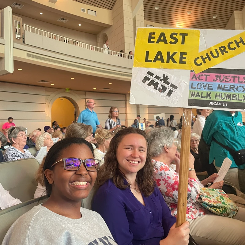 Two smiling young women sit in a crowded auditorium. One holds a sign that reads 'East Lake Church' and 'Act Justly Love Mercy Walk Humbly'.