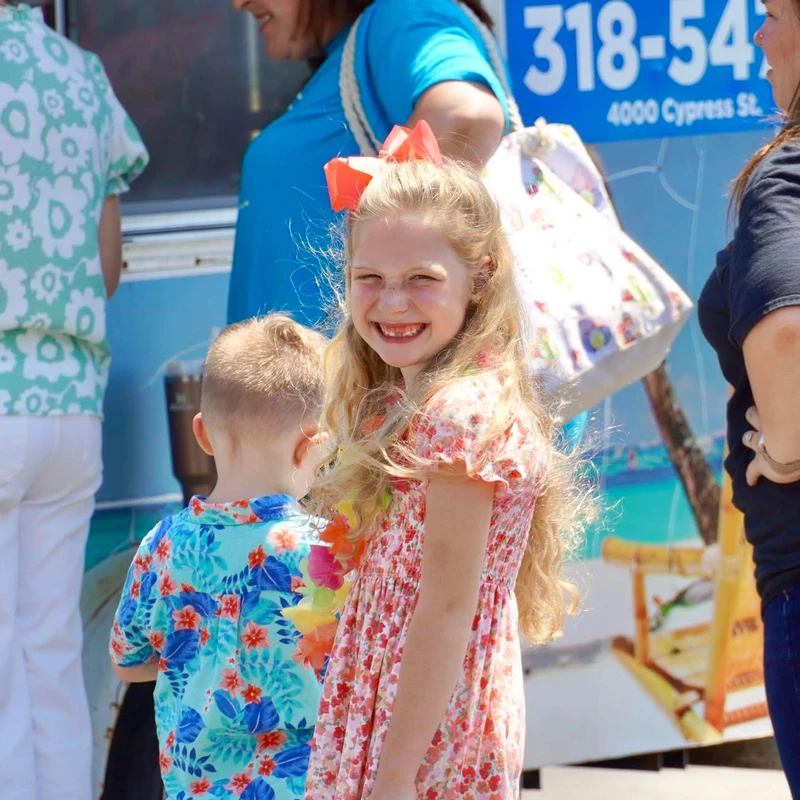 A young girl with long blonde hair and an orange bow smiles at the camera. A little boy in a floral shirt stands beside her in a crowd of people outdoors.