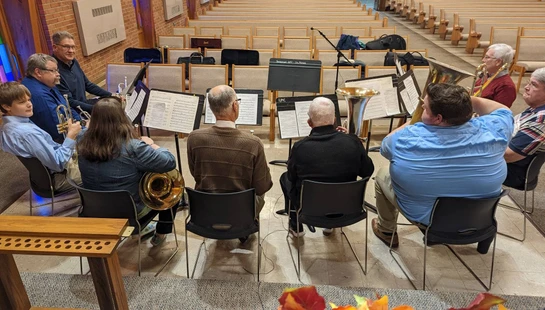 A group of musicians of different ages rehearsing with brass instruments like trumpets and horns in a church sanctuary with empty pews in the background.