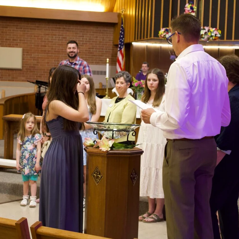 A group of people, including a young woman in a blue dress and a pastor, stand around a baptismal font during a service inside a church sanctuary.