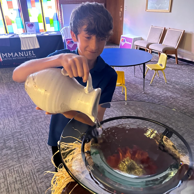 A young boy in a blue shirt pours water from a large white pitcher into a glass font in a church, with a colorful stained-glass window in the background.