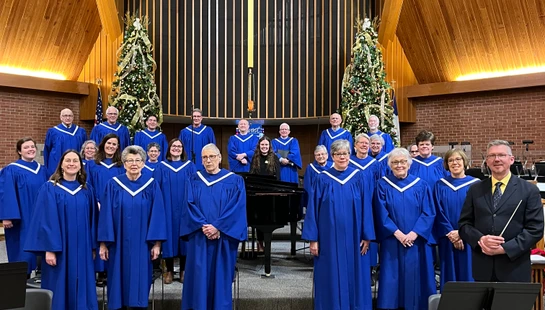 A choir in matching blue robes stands on risers in a church sanctuary, posing for a group photo with their conductor in front of a large cross.