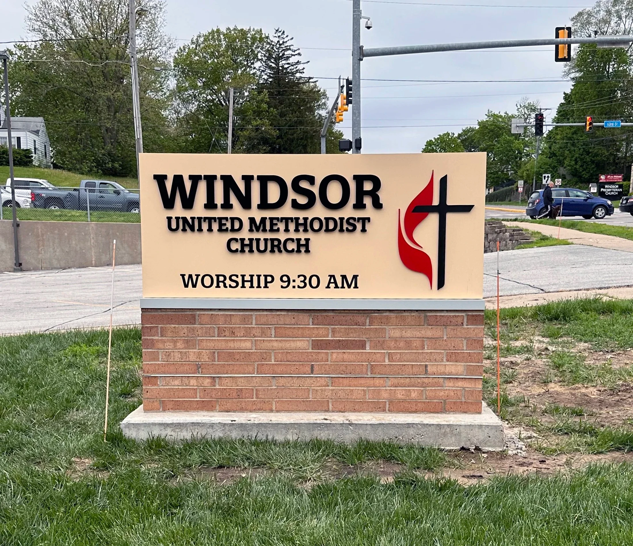 A tan sign on a brick base for Windsor United Methodist Church. The sign shows the worship time as 9:30 AM and features the cross and flame logo.