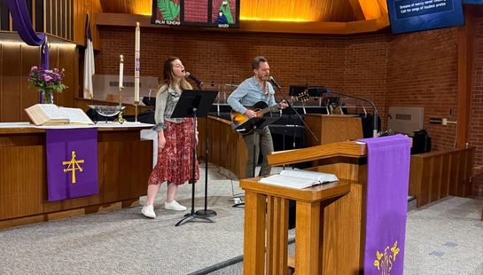 A man playing an acoustic guitar and a woman sing together on a stage in a church sanctuary, leading a worship service during the Lenten season.