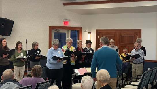 A mixed-age choir, mostly seniors, stands and sings from songbooks for a seated audience in a brightly lit room.