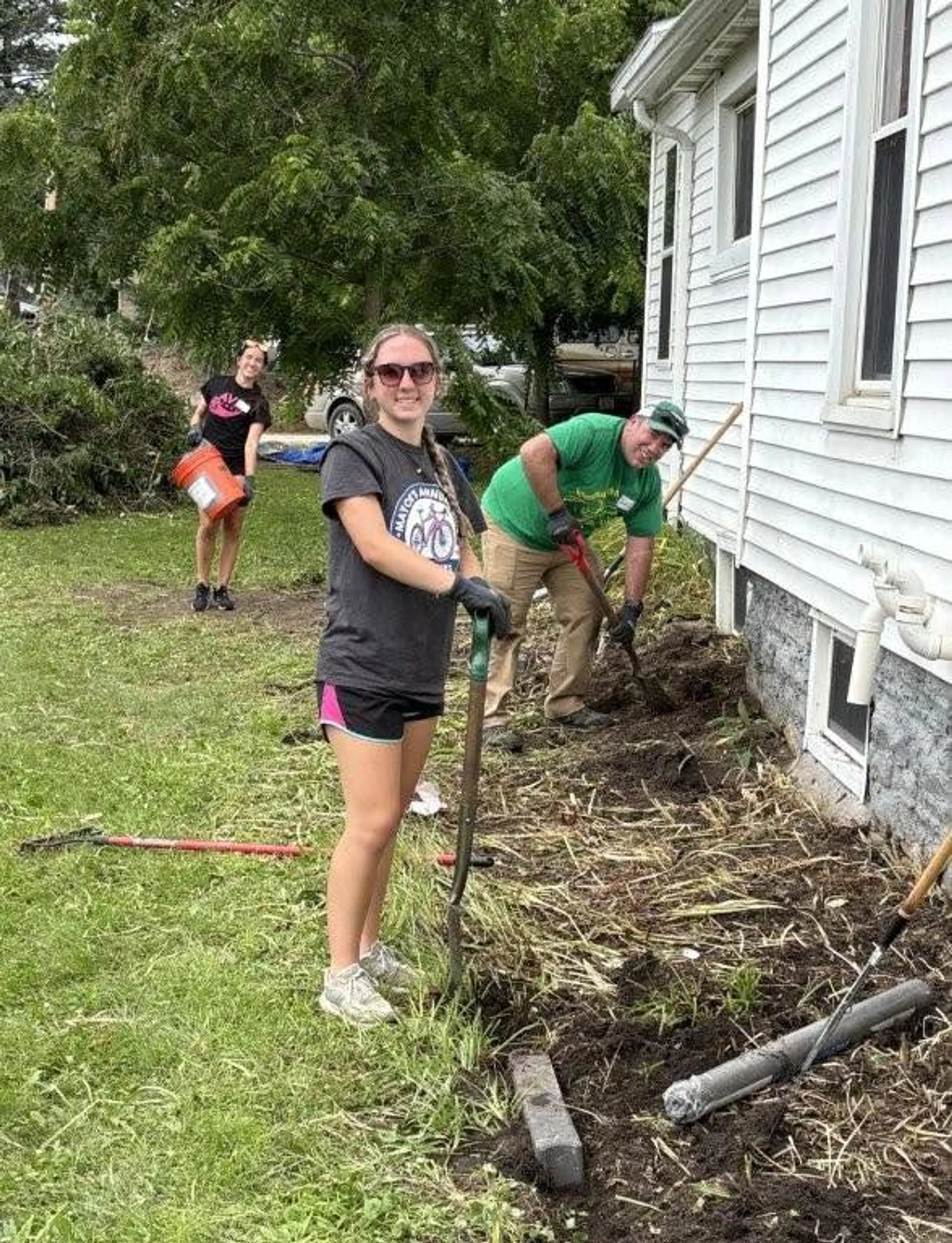 Three volunteers smile while doing yard work outside a house. A young woman holds a shovel, a man digs beside her, and another woman carries a bucket.