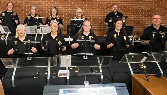A group of ten musicians in matching black shirts smile for a photo behind their music stands, each holding shiny brass handbells against a brick wall.