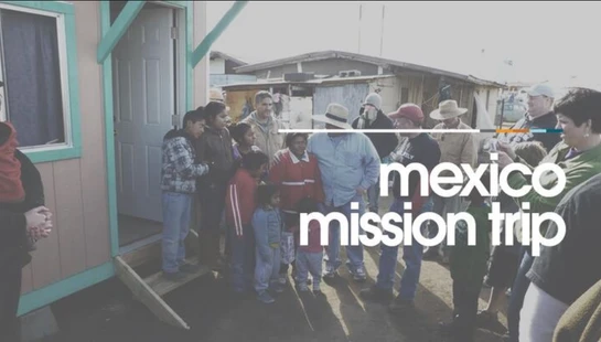 A diverse group of volunteers and local community members stand together outside a simple house during a mission trip in Mexico.