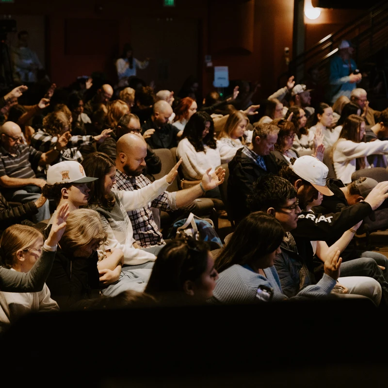 A diverse congregation sits in a dimly lit auditorium, with many people raising their hands in an act of worship or prayer during a service.