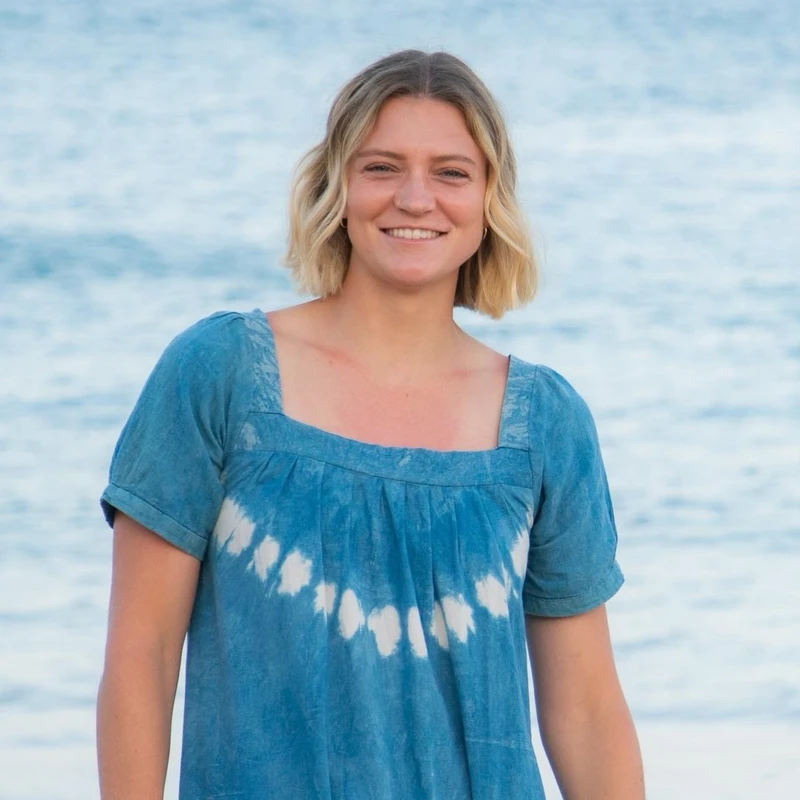 A portrait of a young woman with short blonde hair, smiling at the camera. She is wearing a blue tie-dye dress and standing in front of the ocean.
