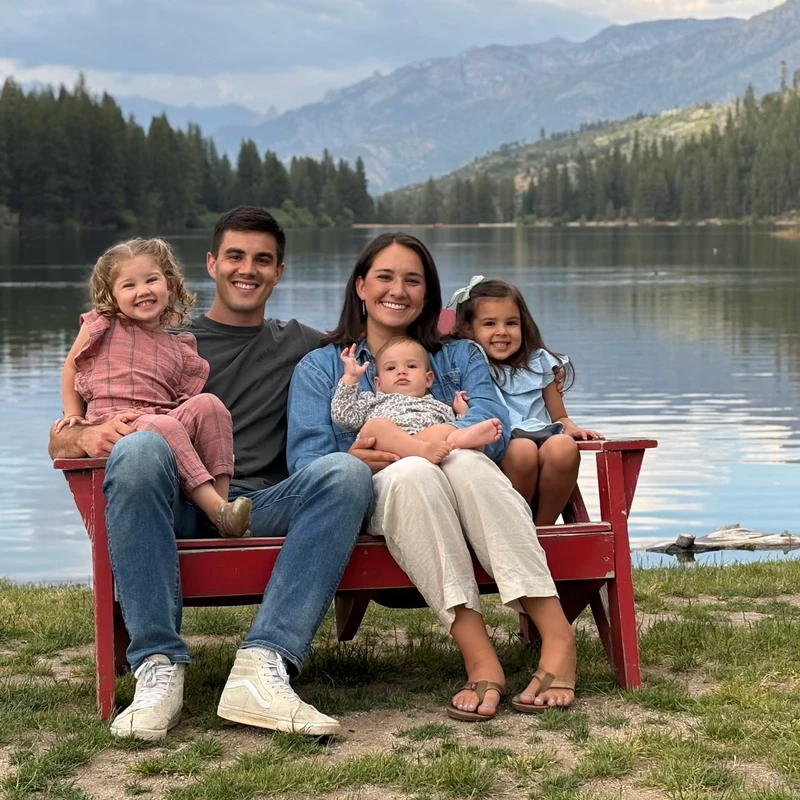 A family of five sits on a red bench by a lake, smiling for a photo. In the background are pine trees and mountains under a cloudy sky.