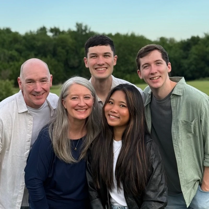 A family of five, with two parents and three young adult children, smile for a group photo in a grassy field with trees in the background.